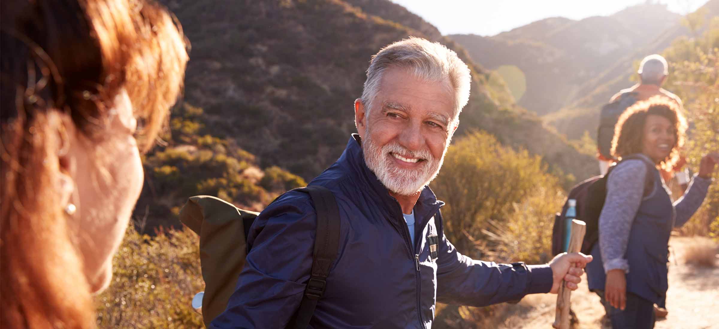 Man helping woman on trail as group of senior friends go hiking in countryside together.