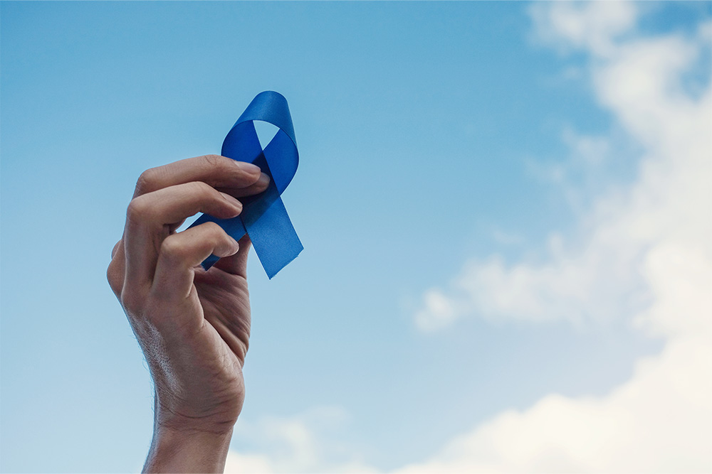 A hand holds a blue prostate cancer awareness ribbon against a blue sky with clouds.